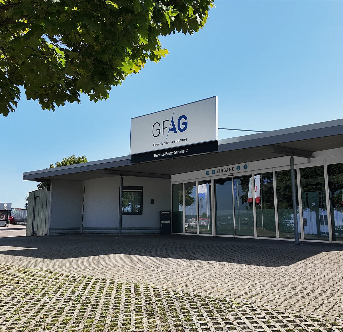 Außenansicht des GFAG-Firmensitzes in Sachsenheim bei klarem, blauem Himmel: Zu sehen ist das ebenerdige Gebäude mit großer Glasfront im Eingangsbereich, dem gepflasterten Vorplatz und einem großen, gut sichtbaren GFAG-Firmenschild auf dem Dach.