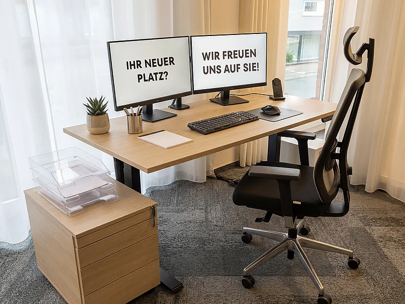 Frei stehender Doppelmonitor-Arbeitsplatz der GFAG in hellem Design: Auf den Monitoren ist eine Begrüßung für potenzielle Bewerber („Ihr neuer Platz? Wir freuen uns auf Sie!“) zu sehen. Der Schreibtisch ist mit Tastatur, Maus, einer kleinen Pflanze und einem Notizblock ausgestattet.