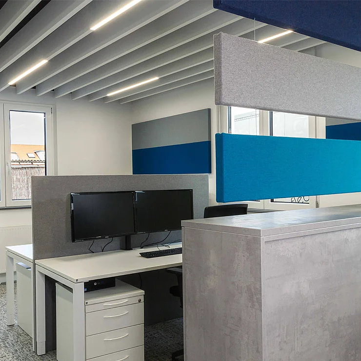 Einblick in das alte Büro der GFAG aus dem Jahr 2017: Doppelarbeitsplatz mit den damals eingesetzten Akustiklösungen. Zu sehen sind graue und blaue Akustik-Baffeln an der Decke mit integrierter Beleuchtung, stoffbezogene Tischtrennwände sowie abgehängte Raumteiler zur Schallabsorption.