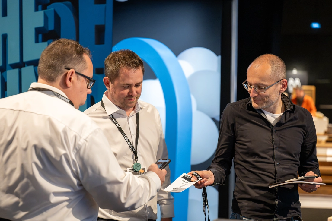 Erfolgreicher Messeauftritt der GFAG auf der Architect@Work in Luxemburg: Mitarbeiter im Gespräch mit einem Messebesucher am Stand. Ein Ticket oder eine Infokarte wird übergeben und zeitgleich mit dem Smartphone erfasst, um wertvolle Kontakte zu knüpfen.