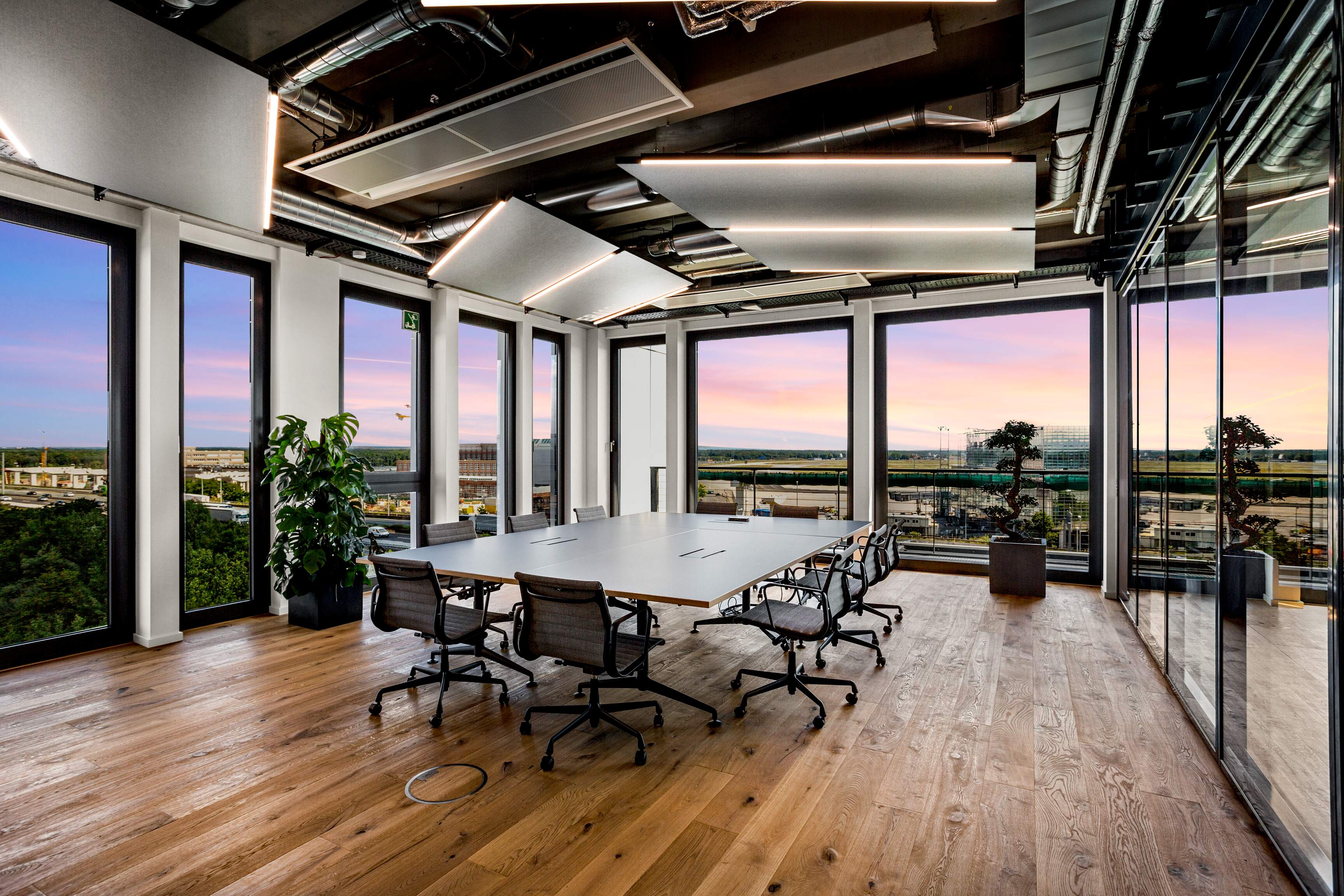 Referenzprojekt der GFAG im Office Gateway Gardens in Frankfurt am Main: Ein moderner Konferenzraum mit edlem Echtholzparkett, umlaufenden Fensterfronten und Abendhimmel-Aussicht. An der offenen Industriedecke sind großformatige, abgehängte Akustiksegel mit integrierten LED-Lampen zur optimalen Schallabsorption und Ausleuchtung installiert.
